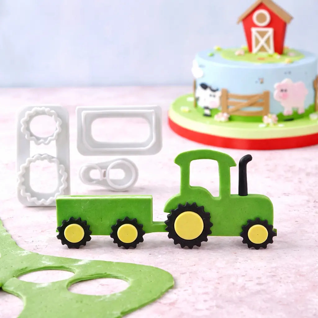 „Mehrteiliges Fondant Ausstecher Set Traktor mit Anhänger – Ausstecher im Fokus mit fertig ausgestochenem Traktor und kleiner Bauernhof-Torte im Hintergrund“